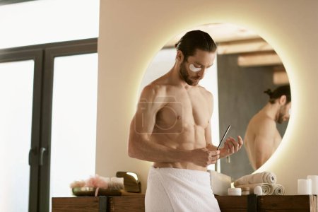 Foto de Un hombre con una toalla mira la lima de uñas mientras hace su rutina de cuidado de la piel matutina en casa.. - Imagen libre de derechos