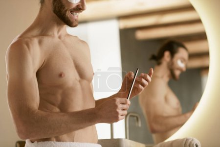Photo for A shirtless man using nail file in front of a mirror at home. - Royalty Free Image