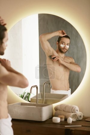 Téléchargez les photos : Un homme avec une serviette se tient devant un miroir, s'engageant dans sa routine matinale de soins de la peau. - en image libre de droit