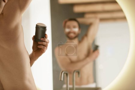 A man using deodorant, surrounded by skincare products, grooming before a mirror.