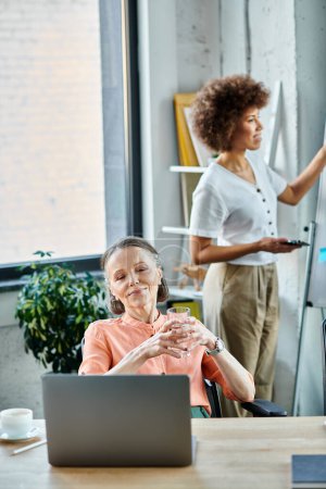 Foto de Mujer alegre centrada en su portátil, inmersa en el trabajo en un entorno de oficina moderno, con su colega afroamericano en el fondo. - Imagen libre de derechos