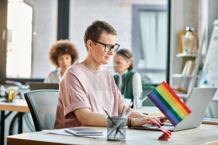 Appealing woman engrossed in work, with a laptop in front of her, with her diverse colleagues on backdrop, pride flag.