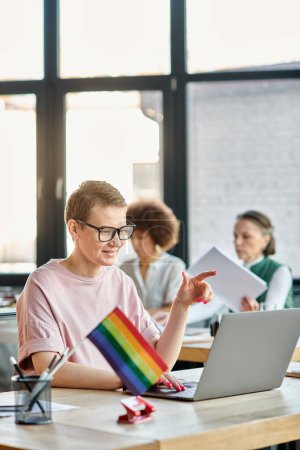 Short haired woman engrossed in work, with a laptop in front of her, with her diverse colleagues on backdrop, pride flag.