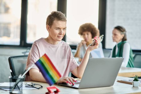 Photo for Pretty woman engrossed in work, with a laptop in front of her, with her diverse colleagues on backdrop, pride flag. - Royalty Free Image