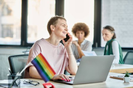 Photo for Jolly woman engrossed in work, with a laptop in front of her, with her diverse colleagues on backdrop, pride flag. - Royalty Free Image