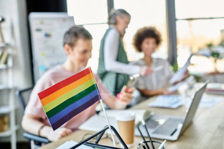 Focus on rainbow flag displayed on office table in front of blurred dverse businesswomen.