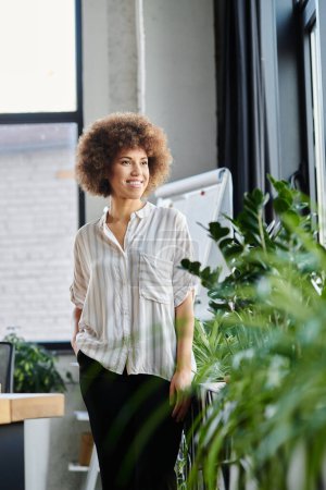 African american young businesswoman stands beside a lush plant in an office setting.