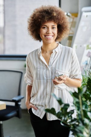 Femme afro-américaine debout en toute confiance dans un cadre de bureau moderne.