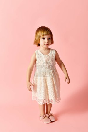 Téléchargez les photos : Petite fille en robe blanche se tient gracieusement sur fond rose. - en image libre de droit