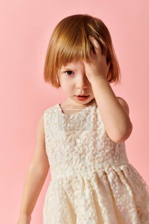 Charming little girl in white dress striking a pose against soft pink backdrop.