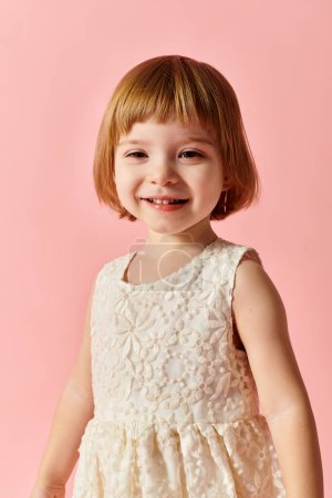 Kleines Mädchen im weißen Kleid posiert vor rosa Hintergrund für ein Porträt.