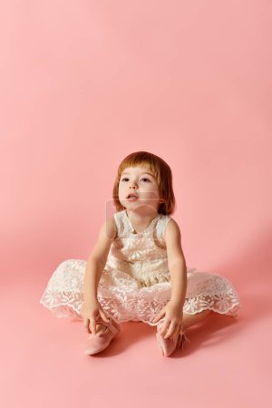 Adorable girl in white dress sitting gracefully on pink backdrop.