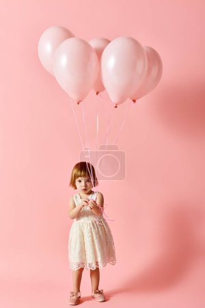 Adorable girl in white dress holding pink balloons on a pink background.