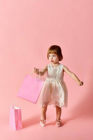 Little girl in white dress holding pink shopping bags on a pink background.