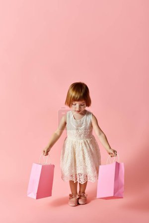 A little girl in a white dress happily holds shopping bags against a pink backdrop.