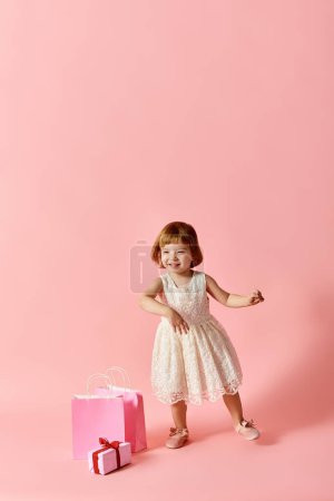 Little girl in white dress with pink gift bags against pink backdrop.