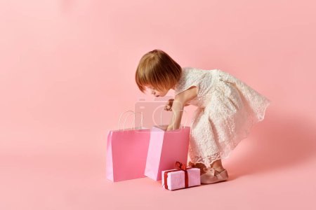 Adorable fille en robe blanche tenant des sacs à provisions roses sur un fond rose.