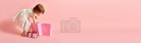 Photo for Adorable girl in white dress holding a vibrant pink shopping bag against a pink background. - Royalty Free Image