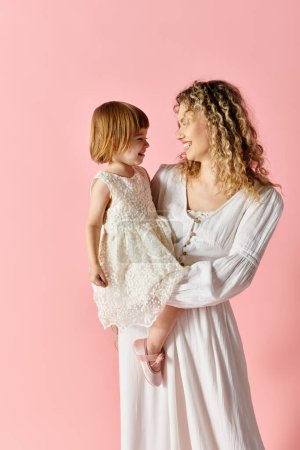 Téléchargez les photos : Mère aux cheveux bouclés et jolie fille en robe blanche debout devant un fond rose vibrant. - en image libre de droit