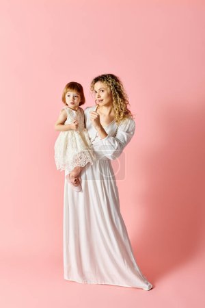 Foto de Una mujer en vestido blanco sostiene a una niña en un vestido blanco sobre un fondo rosa. - Imagen libre de derechos