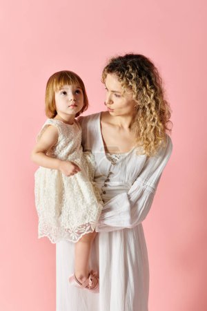 Una madre sosteniendo a su hija con un vestido blanco sobre un fondo rosa.