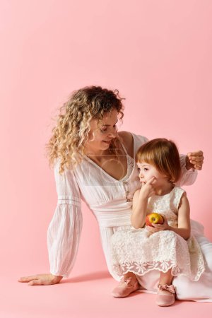 Adorable girl in white dress with curly-haired mom on vibrant pink backdrop.