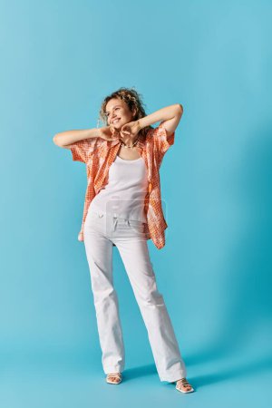 Elegant lady with curly hair stands confidently in white pants and orange shirt on vibrant blue background.