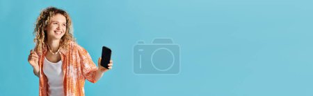 Photo for Woman with curly hair holding cellphone against vibrant blue backdrop. - Royalty Free Image