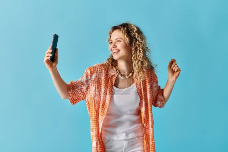 Téléchargez les photos : Femme élégante avec des cheveux bouclés tenant le téléphone sur un fond bleu vibrant. - en image libre de droit