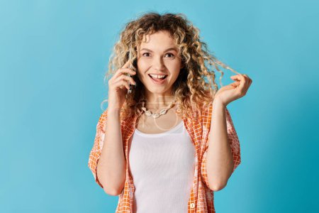 Eine junge Frau mit lockigem Haar spricht auf ihrem Handy vor einem leuchtend blauen Hintergrund.