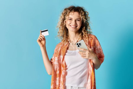 Young woman with curly hair holding credit card and cell phone against colorful backdrop.
