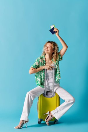 Téléchargez les photos : Stylish woman with curly hair sitting on vibrant yellow suitcase, holding passport. - en image libre de droit