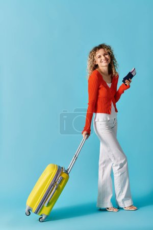 Stylish woman with curly hair holding yellow suitcase and passport and ticket against colorful backdrop.