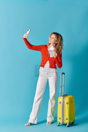 Curly-haired woman taking a selfie with a yellow suitcase against a blue background.