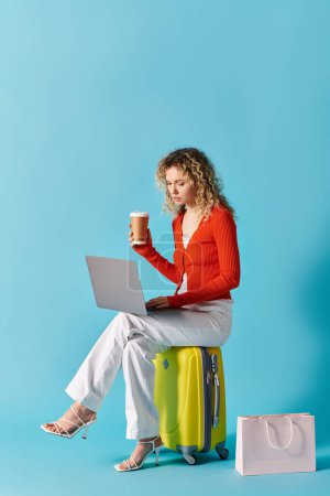 Foto de Una mujer con el pelo rizado se sienta en una maleta, bebiendo café y trabajando en un ordenador portátil. - Imagen libre de derechos