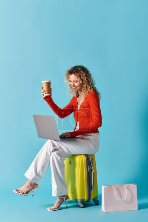 Femme aux cheveux bouclés assis sur une valise avec café et ordinateur portable.