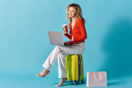 Photo for Curly-haired woman sitting on suitcase, typing on laptop, and enjoying coffee. - Royalty Free Image
