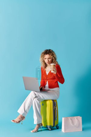 Photo for Curly-haired woman sits on suitcase, sipping coffee, using laptop. - Royalty Free Image