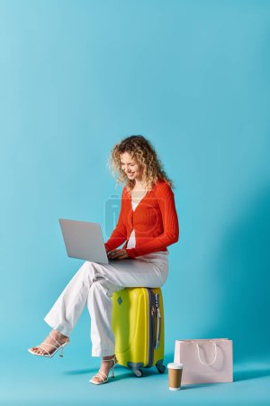 Curly-haired woman sitting on suitcase, using laptop.