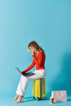 A woman with curly hair sits on a suitcase, working on a laptop.