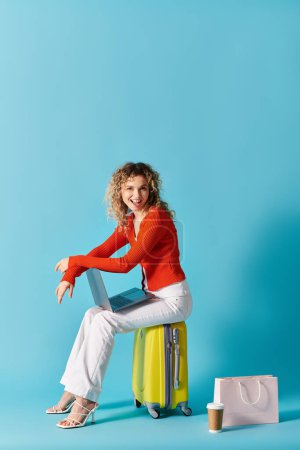 Stylish woman with curly hair sitting on suitcase, working on laptop.