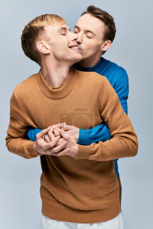 Two men in sweaters affectionately kissing on gray backdrop.