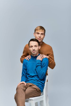 Zwei liebende schwule Männer in legerer Kleidung posieren für ein Foto auf einem weißen Stuhl vor grauem Hintergrund.
