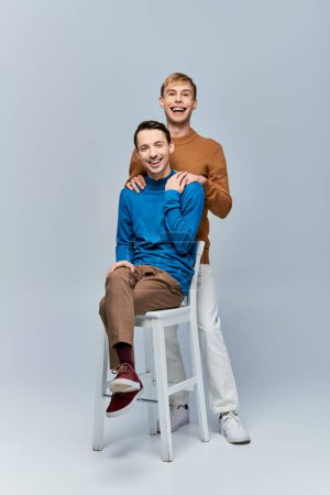 Foto de Dos hombres, una pareja gay amorosa con atuendo casual, se sientan en un taburete posando para la cámara contra un fondo gris. - Imagen libre de derechos