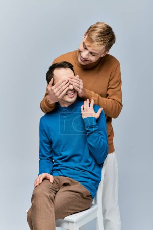 Man in chair, hands of other man covering his eyes.
