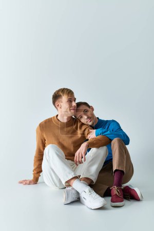 Dos hombres en atuendos casuales sentados en el suelo con los brazos alrededor el uno del otro en el fondo gris.