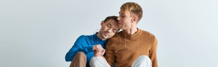 Photo for Two men in casual attires, one holding his leg up, share a moment of intimacy on a gray backdrop. - Royalty Free Image