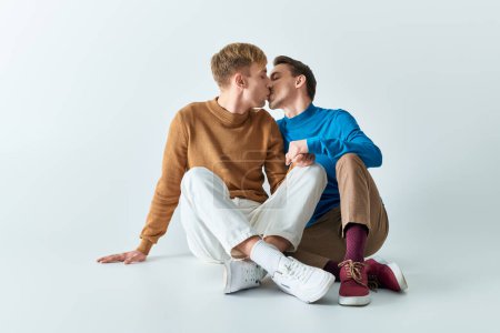 Two young men in casual clothes sitting on the ground kissing each other.