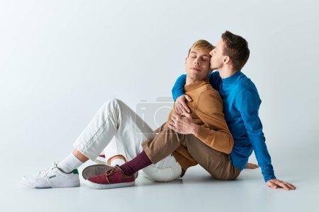 Ein liebevolles schwules Paar sitzt in lässiger Kleidung vor grauem Hintergrund auf dem Boden.