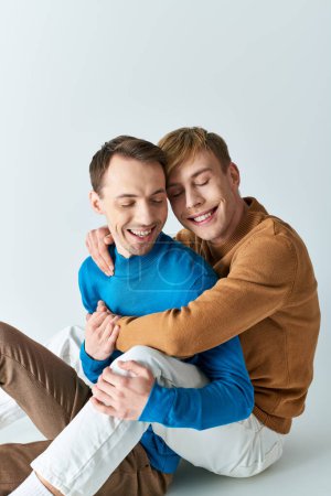 Two men in casual attire sit on the ground, hugging each other tightly.